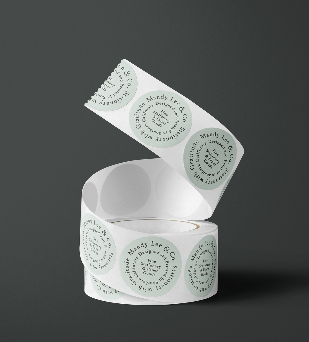 A roll of white paper labels or tape with repeated circular branding printed in pale green, shown on a dark gray studio background. The visible text includes 'Mandy Lee & Co' and 'Stationery with Gratitude'. Fine Stationery & Paper Goods. Custom branded stickers.