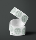 A roll of white paper labels or tape with repeated circular branding printed in pale green, shown on a dark gray studio background. The visible text includes 'Mandy Lee & Co' and 'Stationery with Gratitude'. Fine Stationery & Paper Goods. Custom branded stickers.