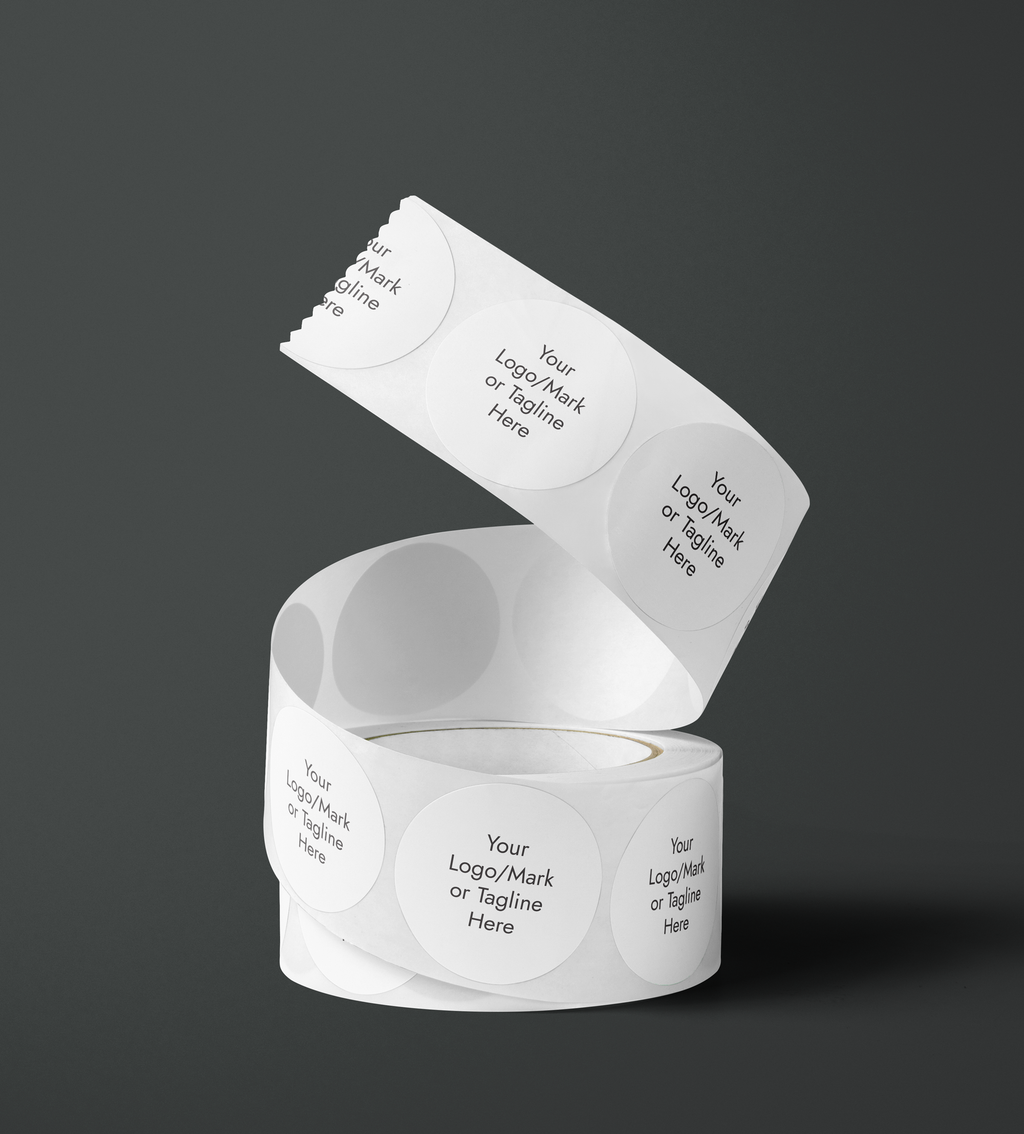 A roll of white paper labels or tape with repeated circular branding printed in black and white shown on a dark gray studio background. The visible text includes 'Your Logo/Mark or Tagline Here'. Fine Stationery & Paper Goods. Custom branded stickers.
